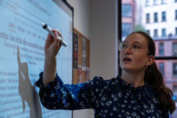 A woman with glasses writing on a whiteboard in a bright classroom setting.