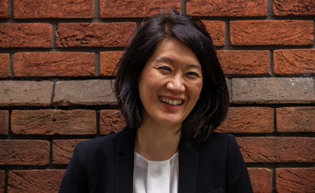 Smiling woman with dark hair in a black blazer against a brick wall background.