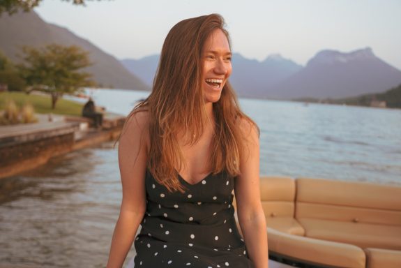 Woman laughing by a lake, wearing a polka dot dress, with mountains in the background.