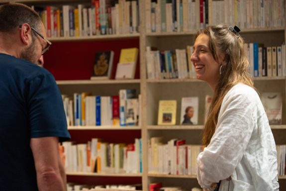 A woman smiling while talking to a man in a library filled with books.