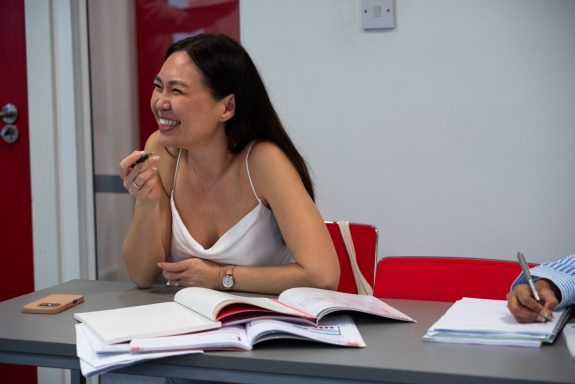 Smiling woman in a white top sits at a table with books, engaged in conversation.